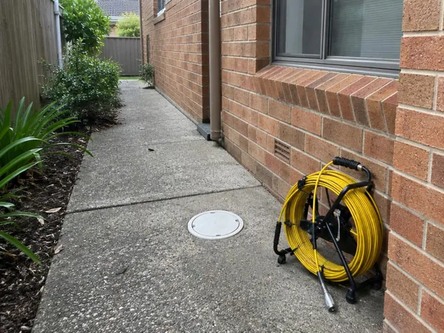 Capped drain inspection opening on a concrete side path beside a brick house and yellow camera reel in Beacon Hill