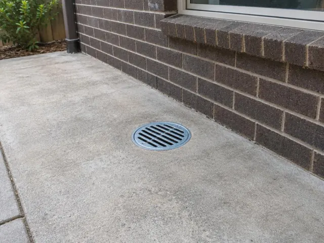Round drainage grate set in dry concrete beside a brick wall in Narrabeen